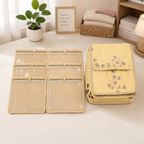 A beige fabric organizer set displayed on a light tablecloth, featuring a folded set of six clear-front pockets and a matching yellow storage bag with a floral vine print. The background shows a small potted plant, a framed sign, and a basket of folded towels.